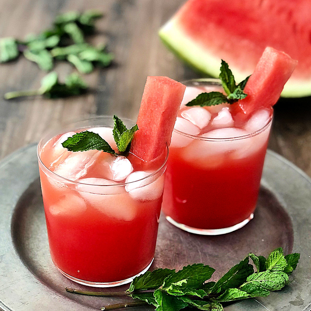 2 glasses of Watermelon Agua Fresca on a silver plate with mint leaves scattered around them.