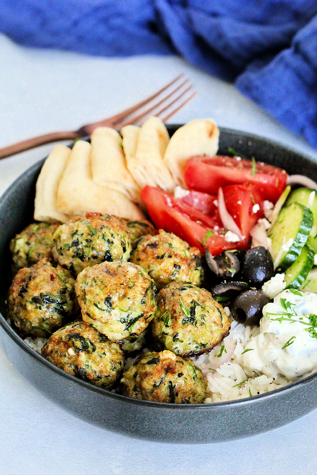 Greek chicken meatballs in gray bowl with tzatziki, tomatoes, cucumbers, olives, feta and pita wedges.