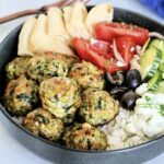Greek chicken meatballs in gray bowl with tzatziki, tomatoes, cucumbers, olives, feta and pita wedges.