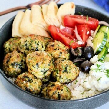 Greek chicken meatballs in gray bowl with tzatziki, tomatoes, cucumbers, olives, feta and pita wedges.