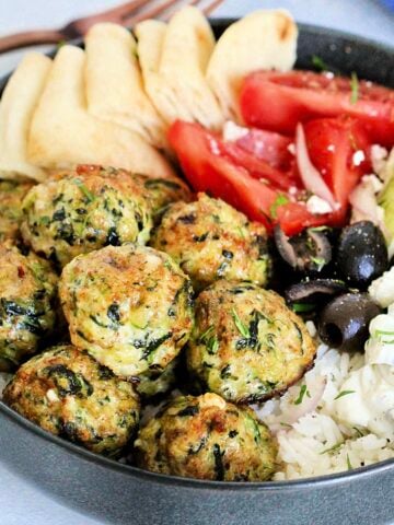 Greek chicken meatballs in gray bowl with tzatziki, tomatoes, cucumbers, olives, feta and pita wedges.
