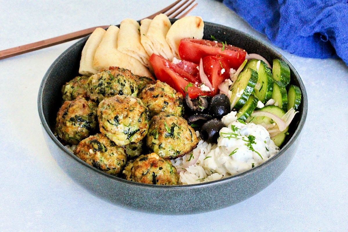 Greek chicken meatballs in gray bowl with tzatziki, tomatoes, cucumbers, olives, feta and pita wedges.