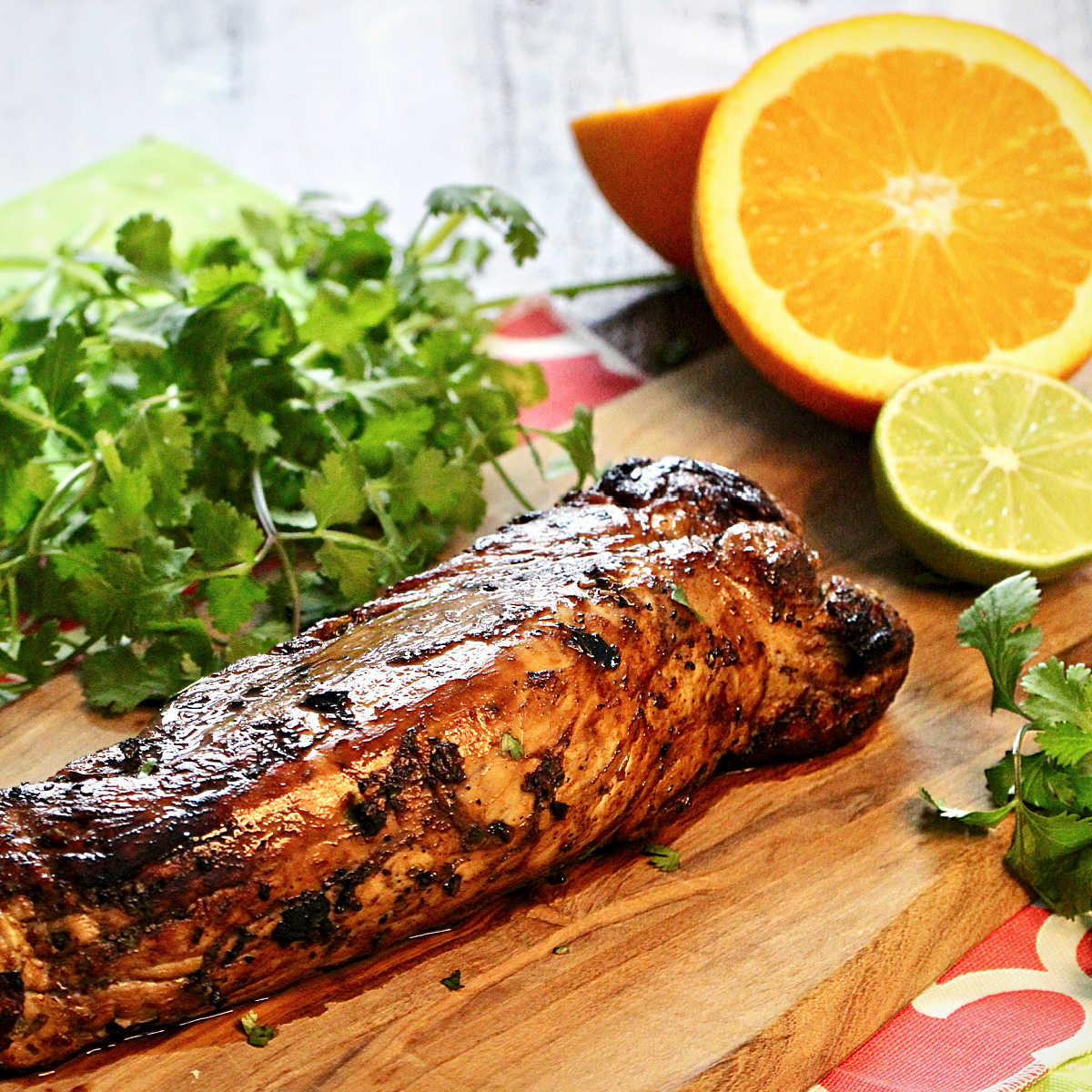 Cuban mojo pork tenderloin on a wooden board with cilantro, an orange and a lime in the background.