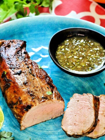 Cuban mojo pork tenderloin on a blue plate with mojo sauce in a black bowl.