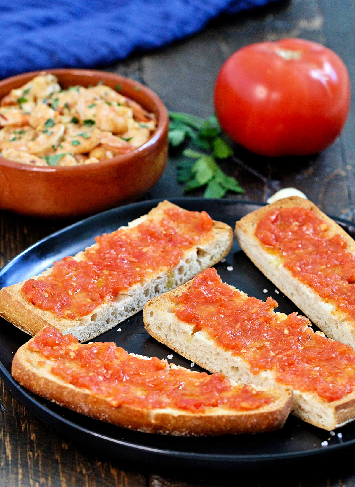 Pan con tomate on a black plate with Spanish garlic shrimp and a tomato in the background.