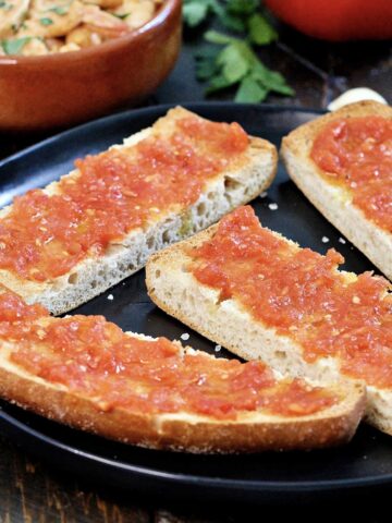 Pan con tomate on a black plate with Spanish garlic shrimp and a tomato in the background.