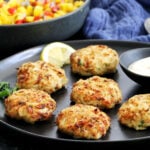 Maryland-style Old Bay crab cakes on a black plate with a bowl of mango salsa in the background.