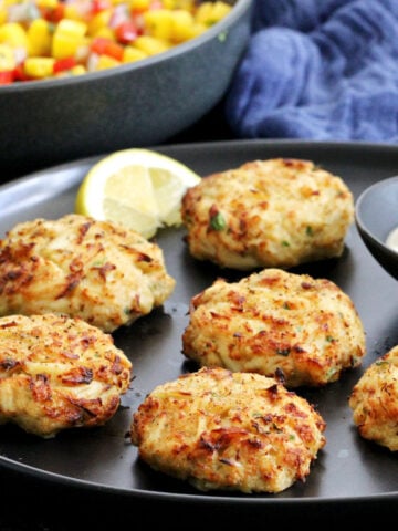 Maryland-style Old Bay crab cakes on a black plate with a bowl of mango salsa in the background.