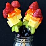 Rainbow fruit kabobs in a mason jar with a black background..