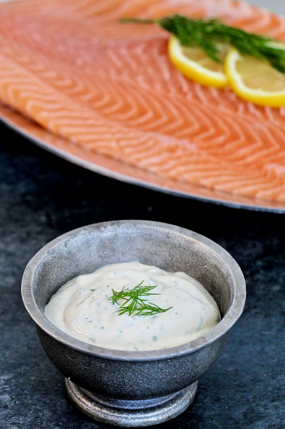 Dill yogurt sauce in a silver bowl with a piece of salmon in the background.