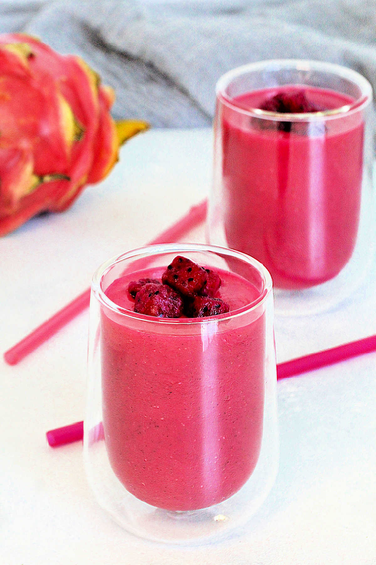 Two dragon fruit smoothies in clear glasses with pink straws and a dragon fruit in the background.