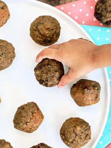 A toddler's hand grabbing a mini banana date muffin from a plate.