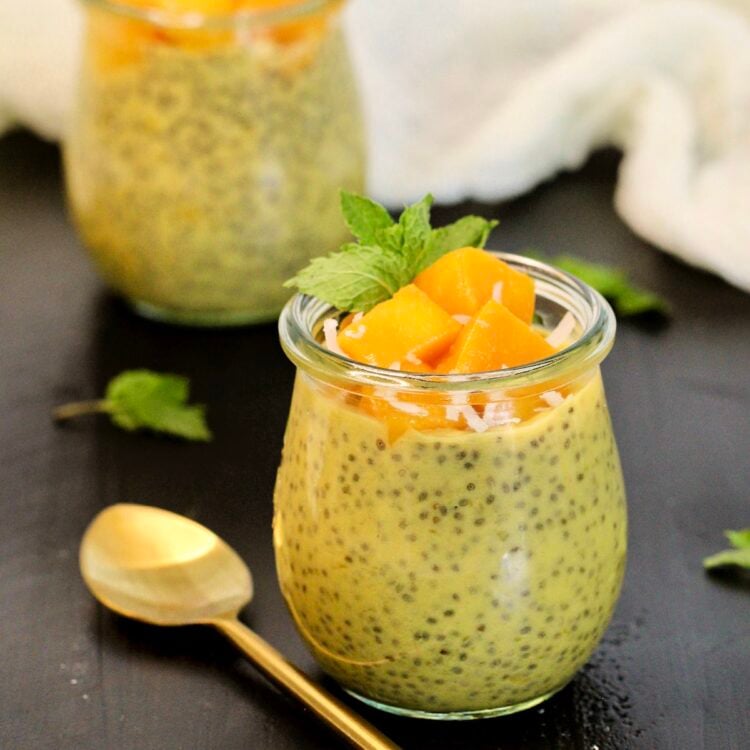 Two mango chia puddings topped with chunks of fresh mango in glass jars with a gold spoon and mint leaves around them.