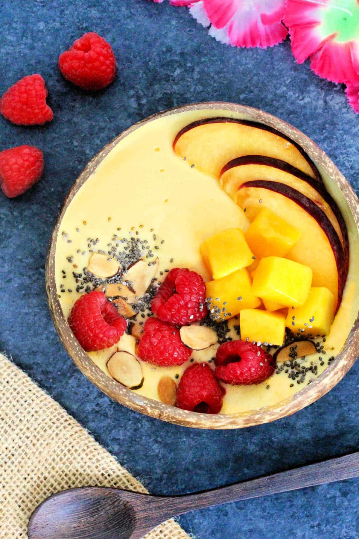 A Mango Smoothie Bowl in a wooden bowl surrounded by a wooden spoon, a cut mango, raspberries and pink flowers.