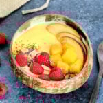 A Mango Smoothie Bowl in a wooden bowl surrounded by a wooden spoon and raspberries.