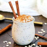 Cinnamon Overnight Oats in a glass jar topped with cinnamon sticks with a gold spoon and oats lying around it.