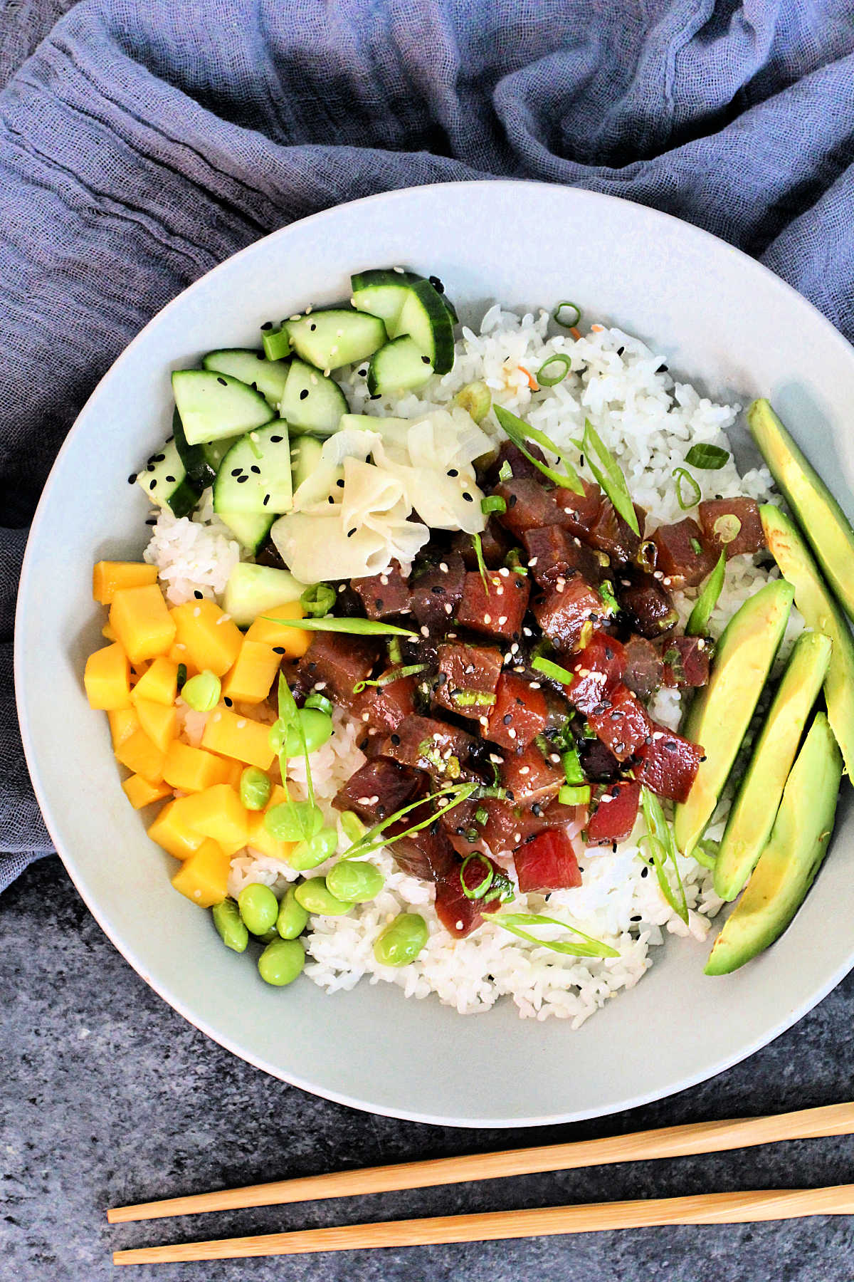 Overhead picture of a Spicy Tuna Poke Bowl in a white bowl with a gray napkin and wooden chopsticks nearby.