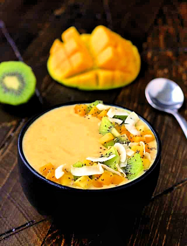 Tropical smoothie bowl in a black bowl with a mango and kiwi in the background.