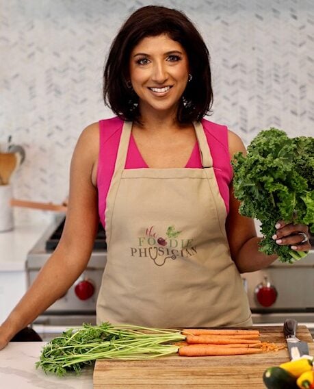 The Foodie Physician standing in a kitchen in an apron with a cutting board.