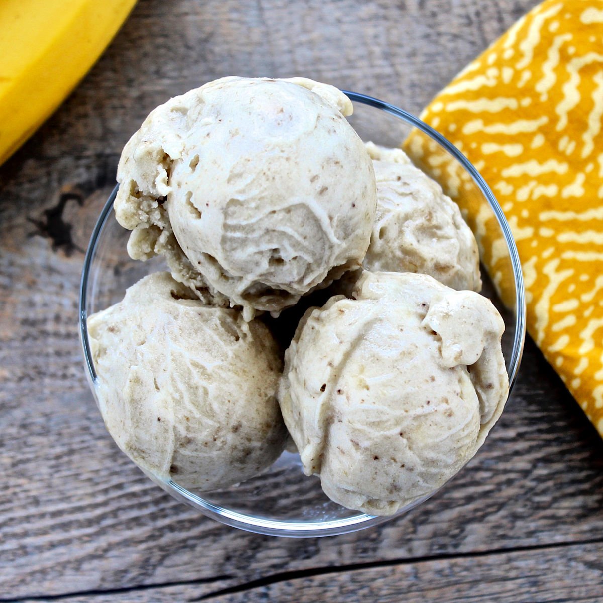 Overhead shot of three scoops of one-ingredient banana ice cream in a glass bowl with bananas and a yellow napkin in the background.