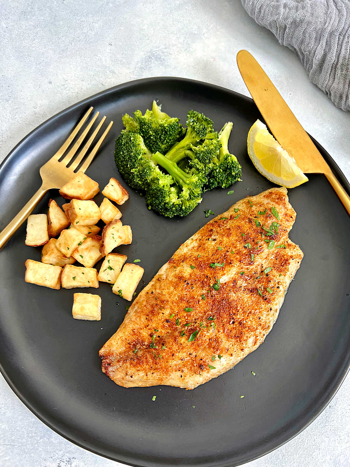 Air fryer tilapia on a black plate with diced potatoes, broccoli and a lemon wedge.
