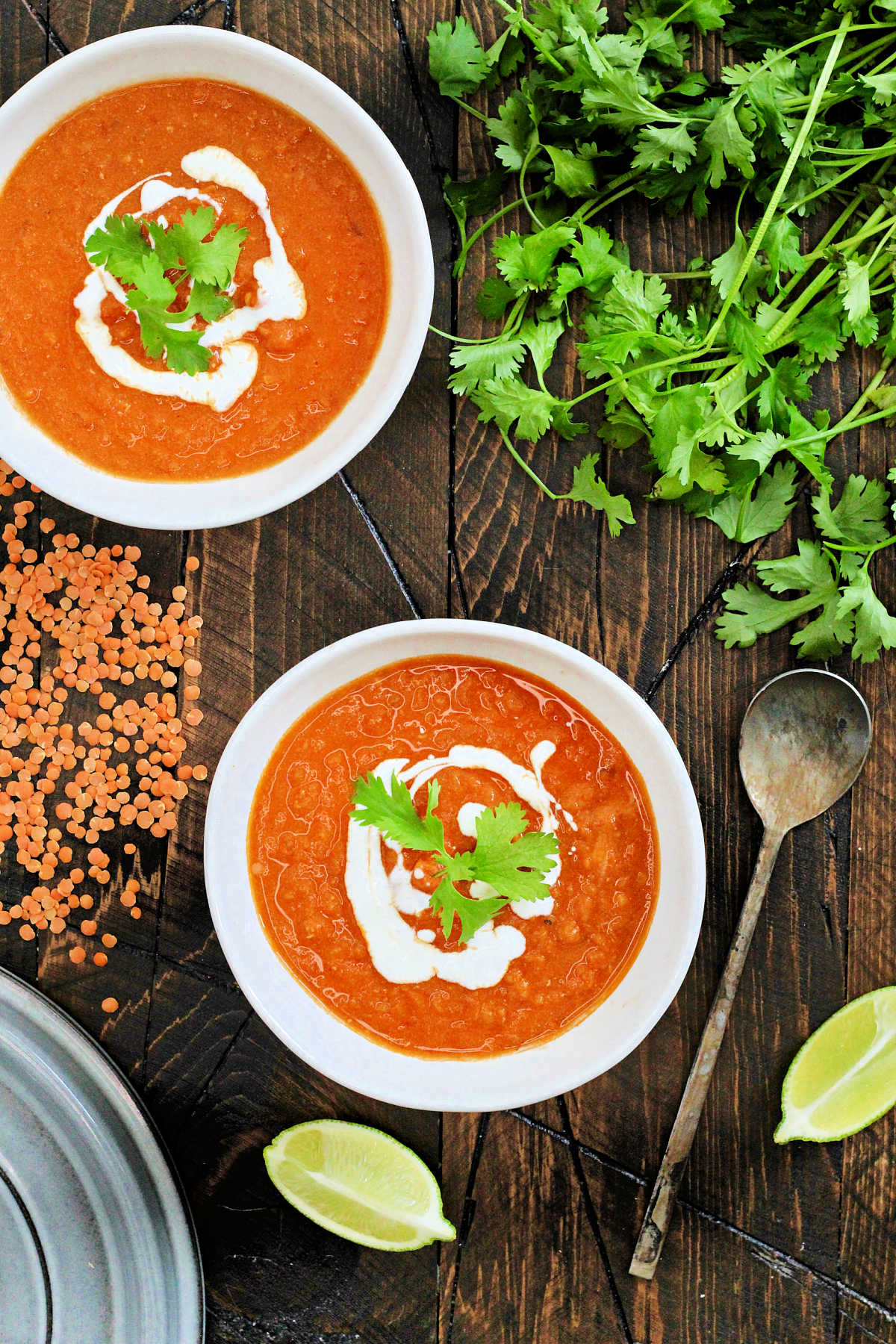 Two bowls of coconut curry lentil soup on a wooden board with a metal spoon, red lentils, and cilantro on a wooden board.