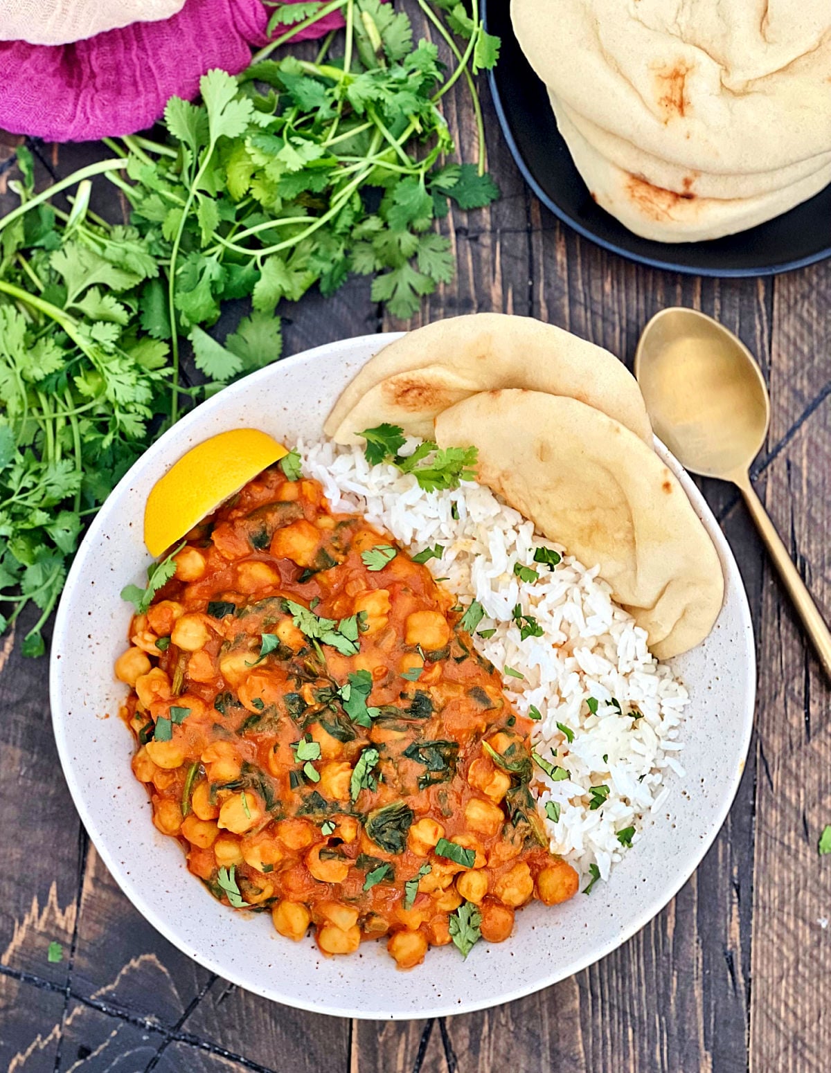 Overhead shot of chickpea spinach curry in a white bowl with basmati rice, naan bread and a bunch of cilantro on a wooden board.
