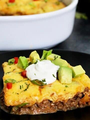 Piece of easy keto taco casserole on a black plate in front of a white casserole dish.