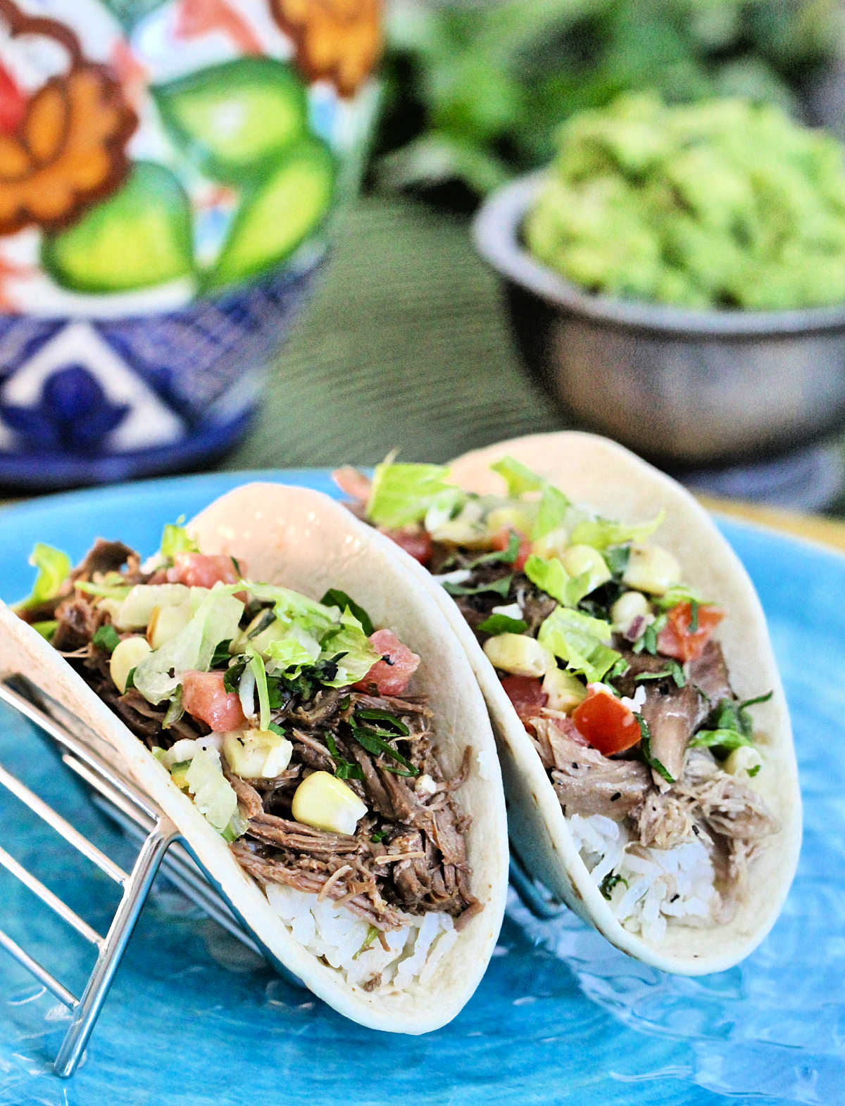 Barbacoa vs. carnitas. One barbacoa taco and one carnitas taco in a metal taco stand with guacamole in background.