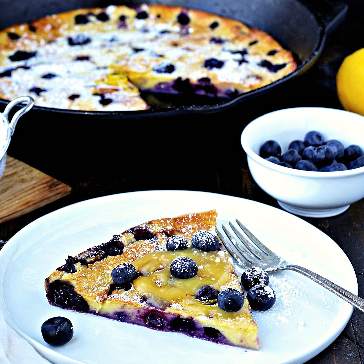 Slice of blueberry lemon dutch baby on a white plate with cast iron skillet in background.