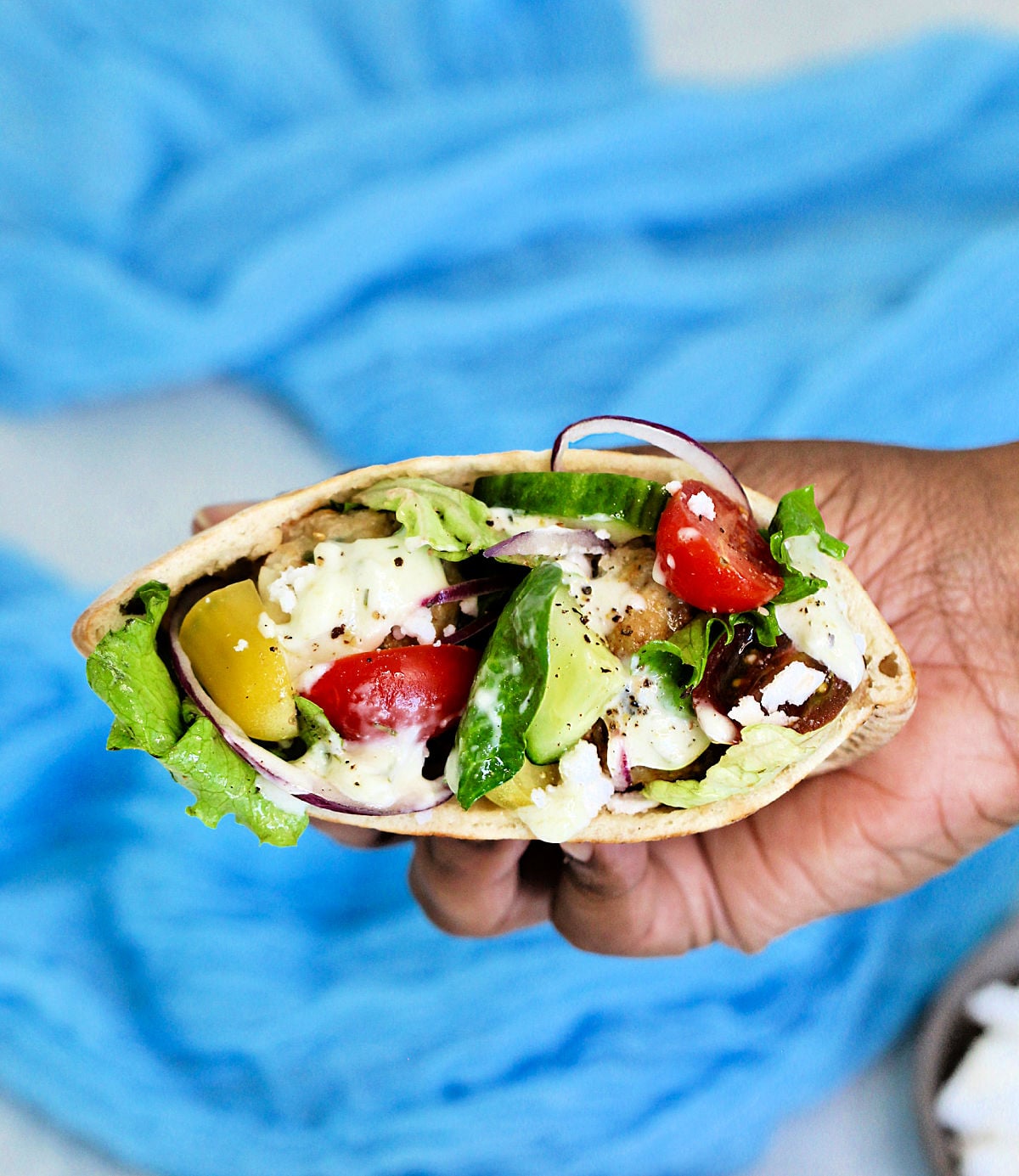 A Greek pita sandwich being held in front of a blue cloth.