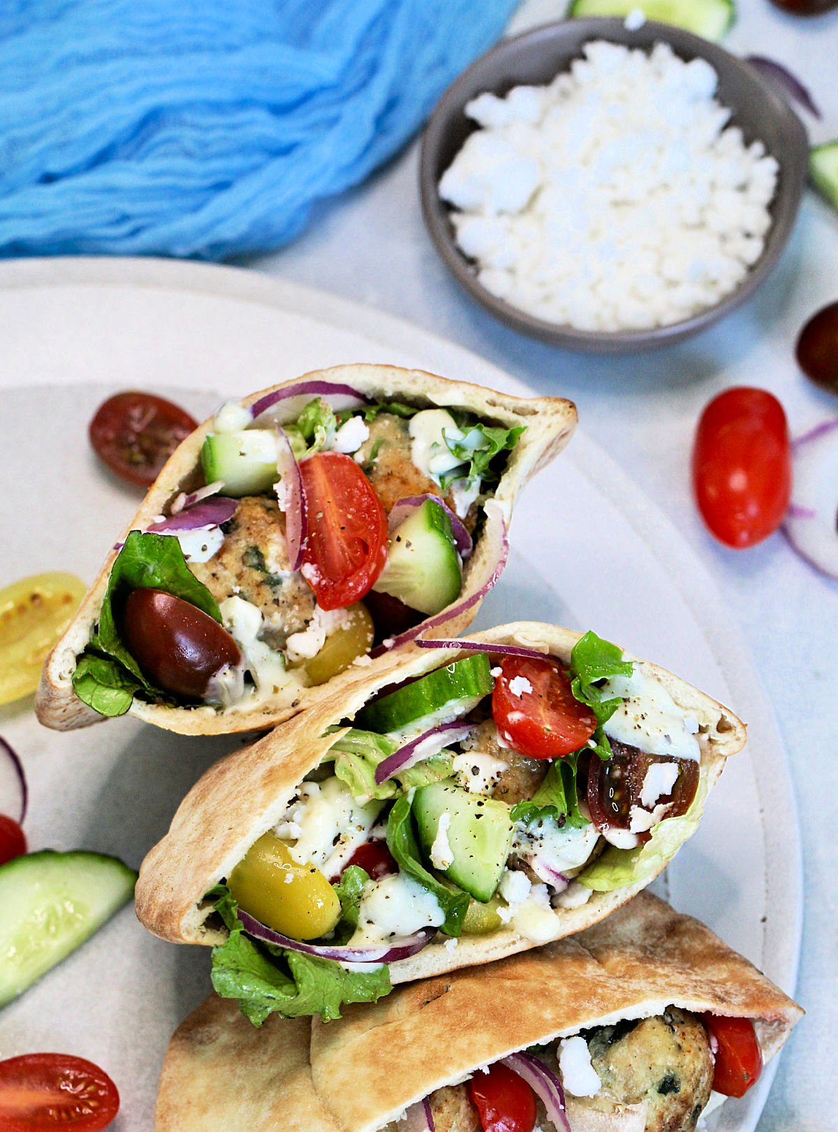 Three Greek pita sandwiches on a round plate with a bowl of feta cheese next to it.