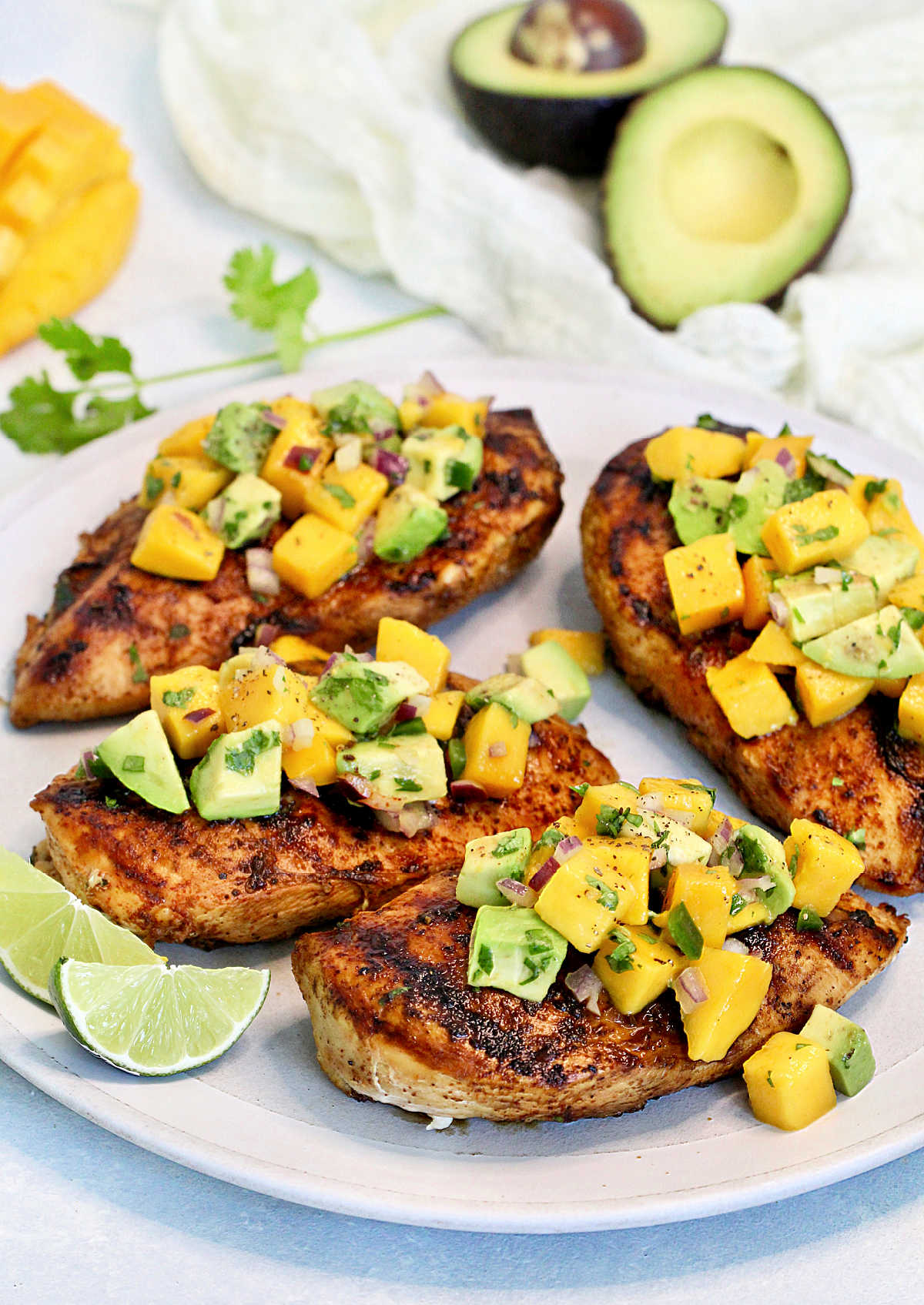 Grilled chicken with mango avocado salsa on a white plate with mango and avocado in the background.