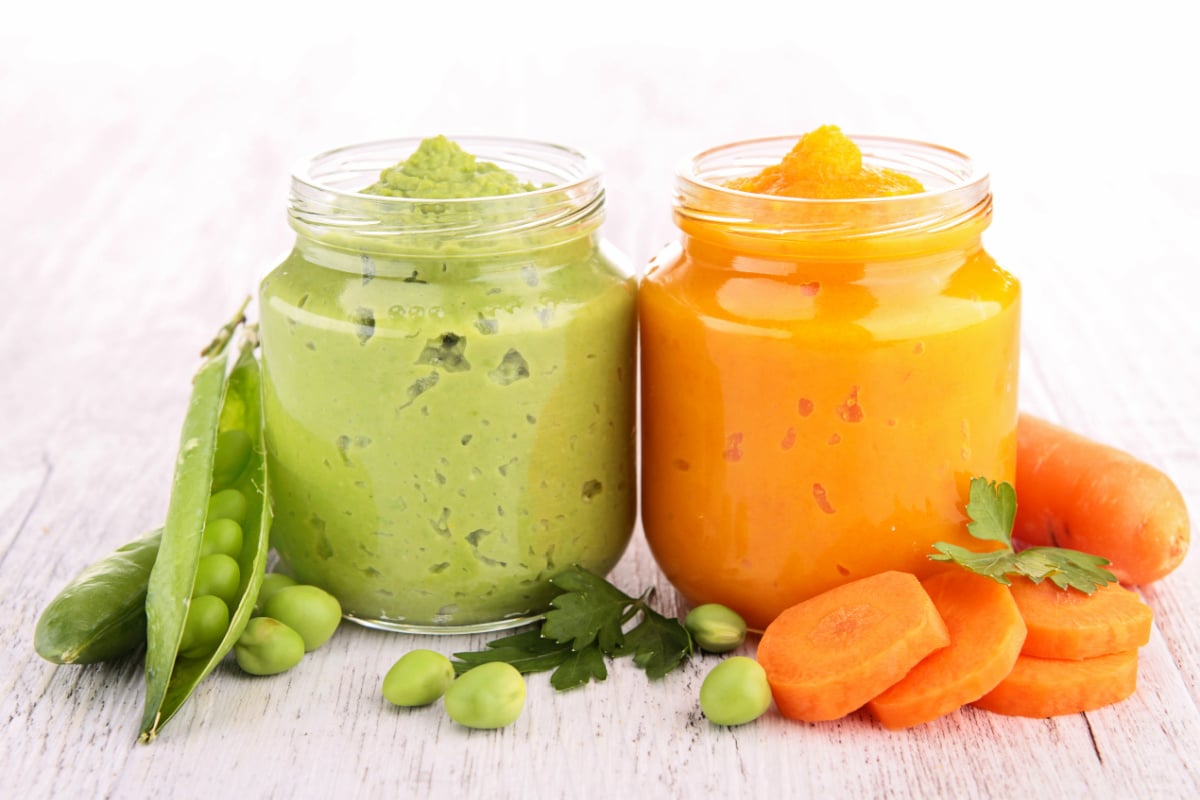 A jar of pea puree and a jar of carrot puree with peas and carrots around them
