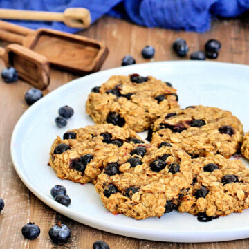 Blueberry oatmeal cookies on white plate surrounded by blueberries, wooden measuring spoons and a blue napkin.