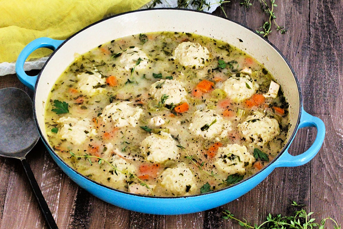 Chicken and Dumplings in a light blue braiser on a wooden board with a metal serving spoon and yellow napkin nearby.