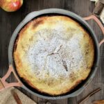 An Apple Dutch Baby in a pan on a wooden board surrounded by apples, cinnamon sticks and measuring spoons.