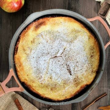 An Apple Dutch Baby in a pan on a wooden board surrounded by apples, cinnamon sticks and measuring spoons.