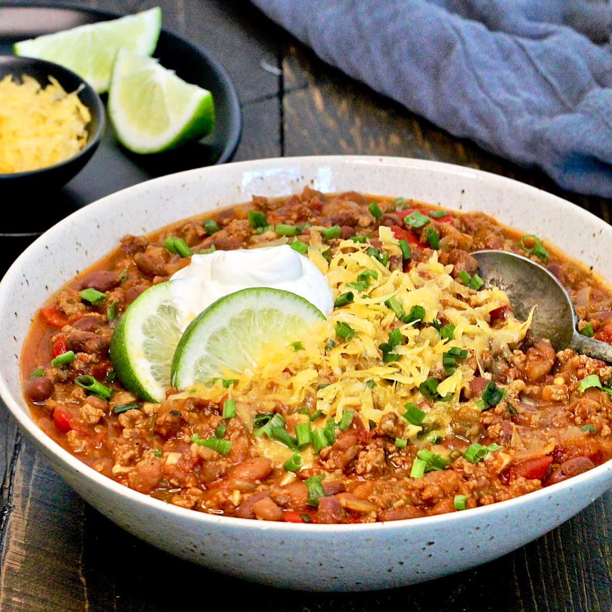 The Ultimate Turkey Chili garnished with cheese, sour cream and lime in a white speckled bowl.