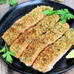 Four pieces of salmon oreganata on a black plate with parsley and lemons.