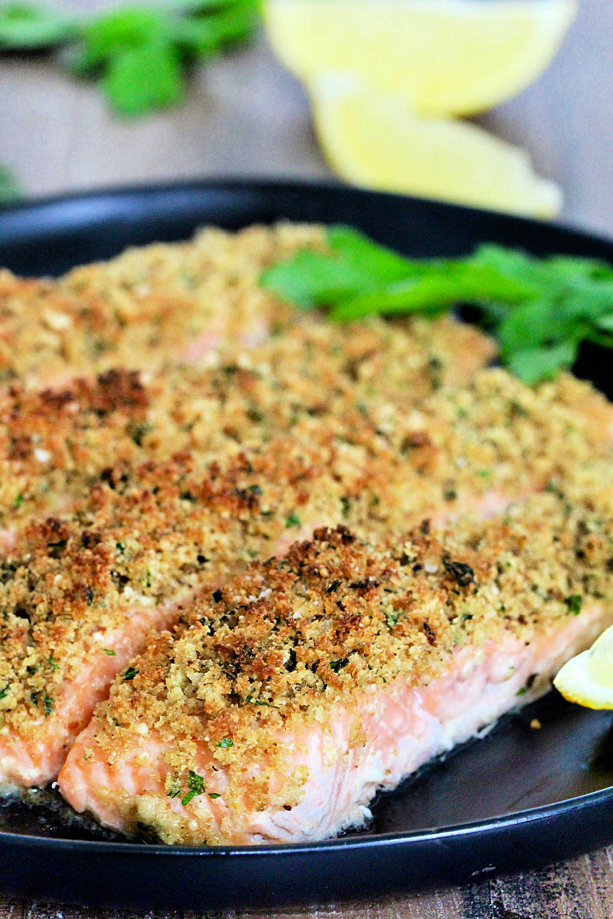 Four pieces of salmon oreganata on a black plate with parsley and lemons.