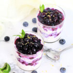 Two glasses of Easy Blueberry Overnight Oats topped with blueberries, lemon zest and mint on a white board with blueberries and a silver spoon around them.