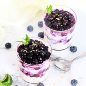 Two glasses of Easy Blueberry Overnight Oats topped with blueberries, lemon zest and mint on a white board with blueberries and a silver spoon around them.
