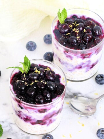 Two glasses of Easy Blueberry Overnight Oats topped with blueberries, lemon zest and mint on a white board with blueberries and a silver spoon around them.