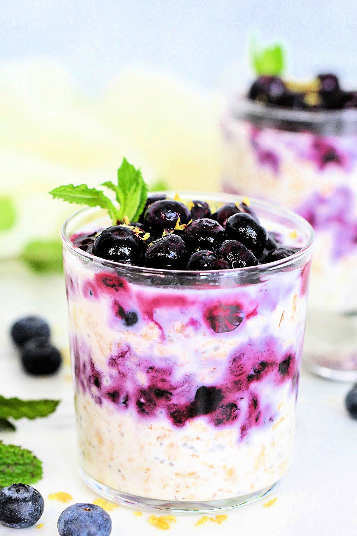 Two glasses of Easy Blueberry Overnight Oats topped with blueberries, lemon zest and mint on a white board with blueberries around them.