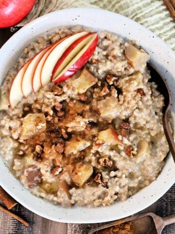 Easy Crockpot Apple Cinnamon Oatmeal in a bowl garnished with apple slices and cinnamon on a wooden board with an apple, cinnamon sticks and measuring spoons around it.