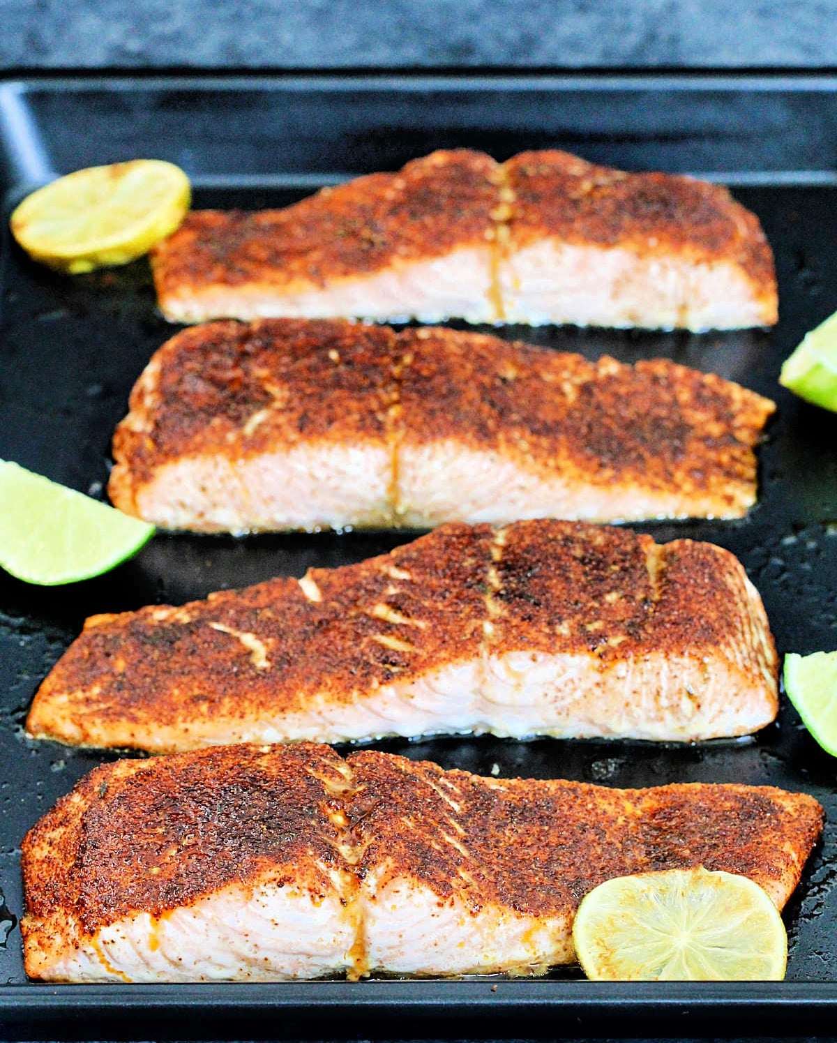 Four fillets of Low Sodium Chile Lime Salmon on a black baking sheet with limes slices around them.