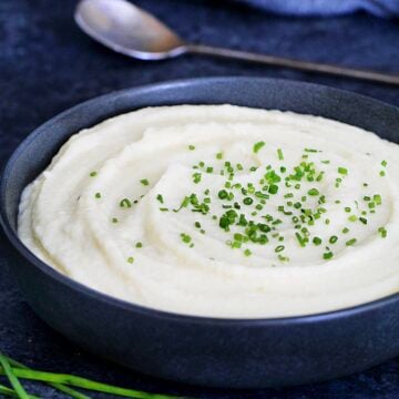 Celeriac (celery root) puree garnished with scallions in a dark bowl with a metal spoon and scallions around it.