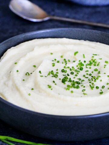 Celeriac (celery root) puree garnished with scallions in a dark bowl with a metal spoon and scallions around it.
