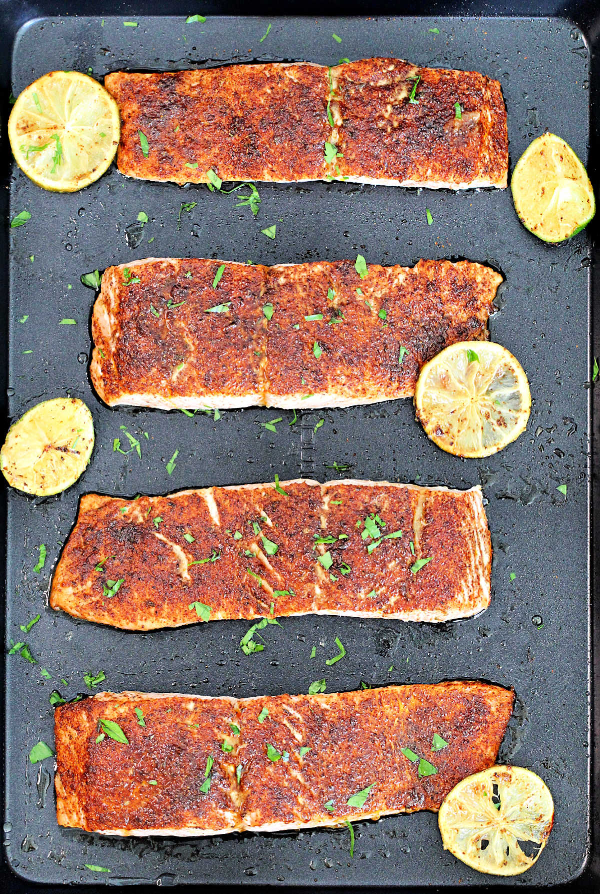 Four fillets of Low Sodium Chile Lime Salmon on a black baking sheet with limes slices around them.